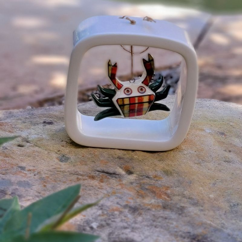 A multi colored plaid crab dangles from an open white ceramic frame sitting on a rock. 